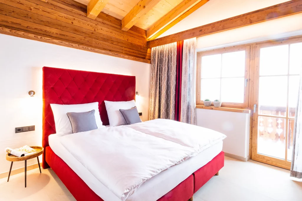 Hochwertiges Doppelbett mit einem eleganten, roten und gesteppten Kopfteil in einem stilvollen Schlafzimmer des Ferienhauses Mandlberggut in Radstadt/Mandling. Das Schlafzimmer bietet eine luxuriöse Einrichtung mit weißen Bettdecken, weichen Kissen und großzügigen Panoramafenstern, durch die viel Tageslicht einfällt und die einen atemberaubenden Blick auf die umliegende Alpenlandschaft ermöglichen.