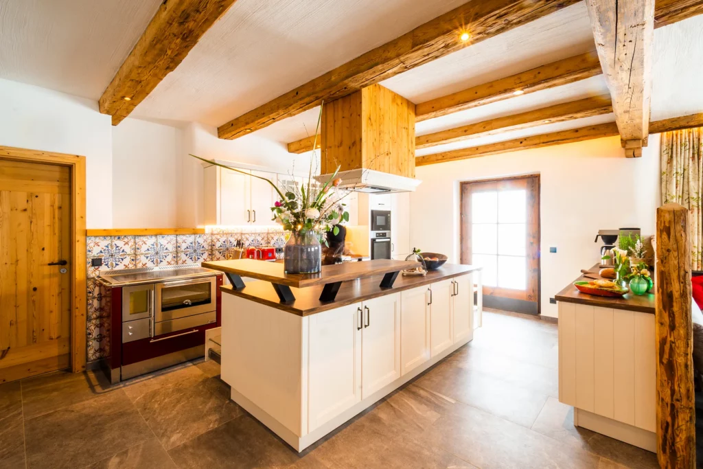 Helle Küche im Ferienhaus Mandlberggut mit rustikalen Holzbalken, moderner Kochinsel, traditionellem Holzofen und dekorativen Fliesen in alpinem Stil.